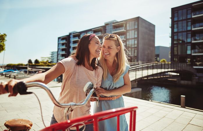 Two close friends laughing while walking with a bicycle along the river walk