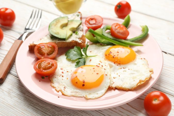 Two fried eggs with tomatoes and avocado toast on pink plate