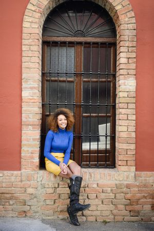 Female with curly hair wearing bright blue shirt and yellow skirt sitting in front of large window