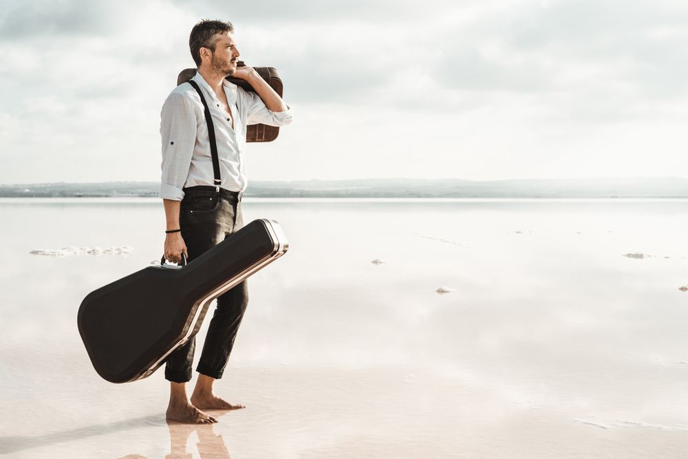 Focused stylish man with suitcase and guitar gig bag on seaside