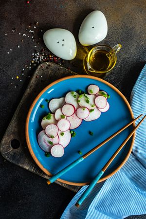Top view of sliced radishes served with chopsticks on blue plate with side of oil and seasoning