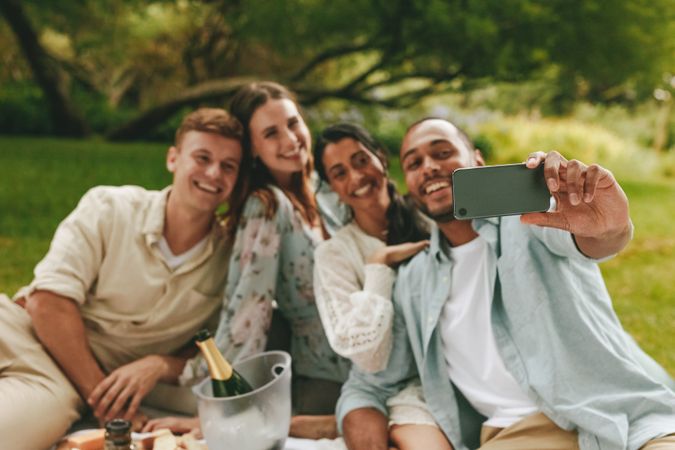 Happy women and men smiling for selfie while sitting on picnic blanket