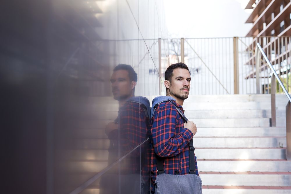 Man in plaid shirt leaning on wall outdoors with a bag on his shoulder on a sunny day