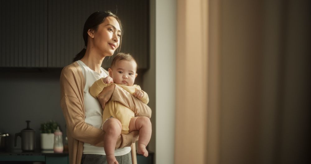 Young Asian Mother Holding her Baby in her Arms While Standing Next to a Window at Home.
