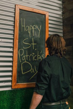 Back view of a man writing "Happy St. Patrick's Day" on wooden framed chalk board