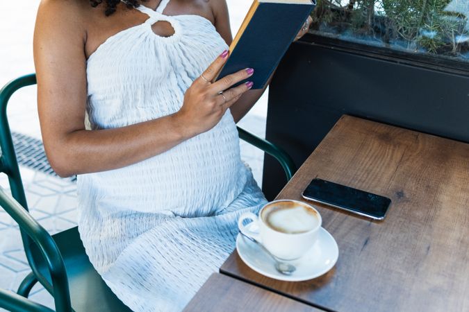 Anonymous pregnant Black woman reading book and drinking coffee