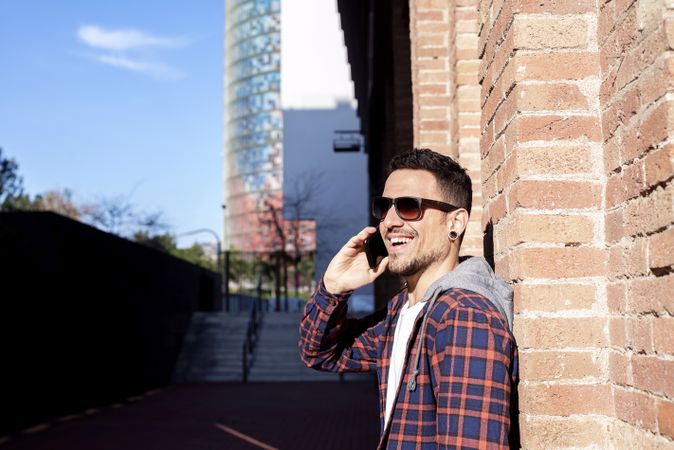 Smiling bearded man leaning on a brick wall wearing sunglasses while using a smartphone