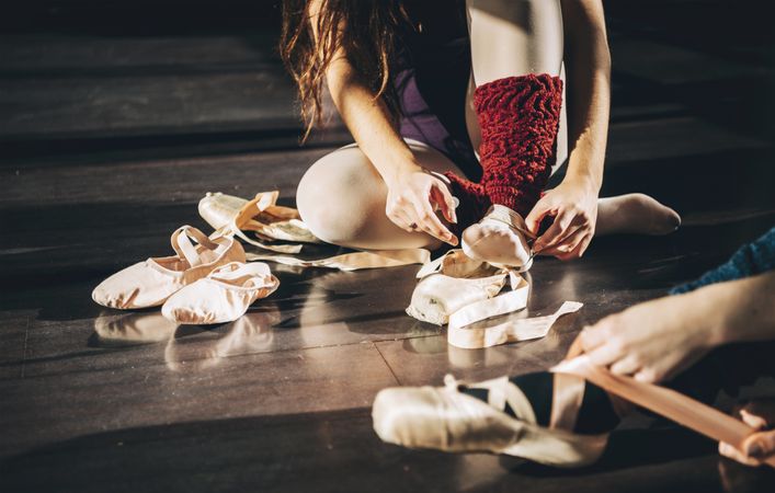 Crop women sitting on wooden floor putting on satin pointe shoes in sunlight.