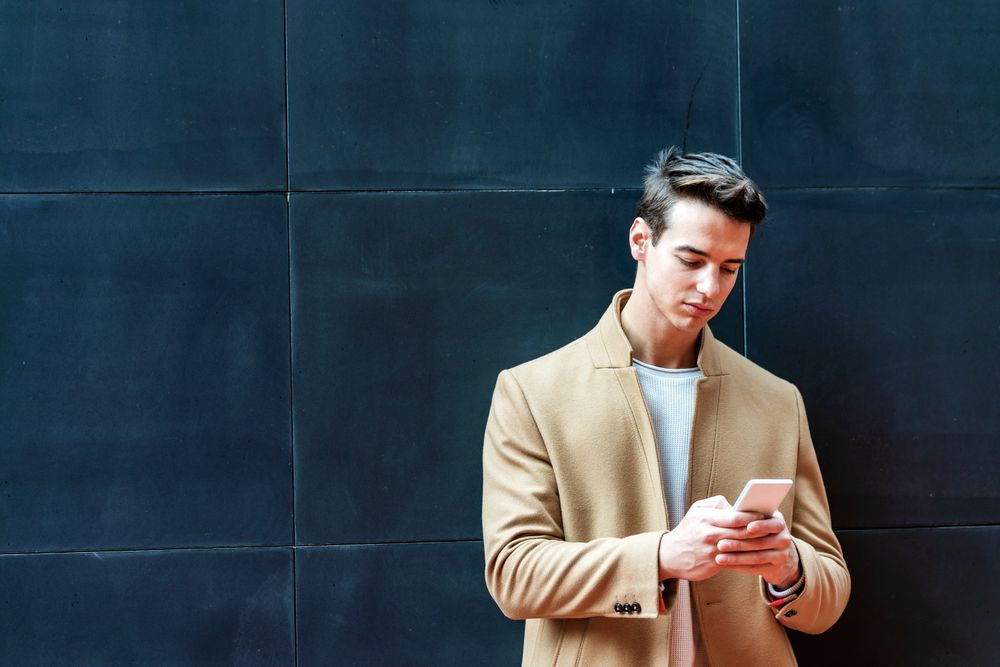 Man in fall coat leaning back on dark outdoor wall while texting