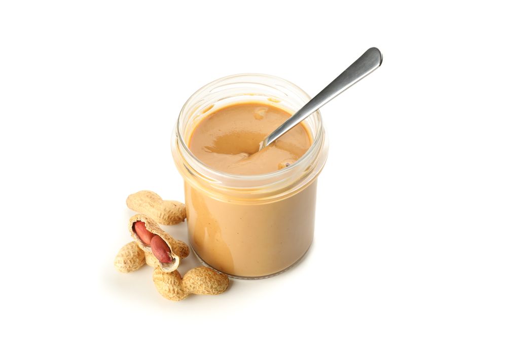 Creamy peanut butter in glass jar, peanut and spoon isolated on light background. A traditional product of American cuisine