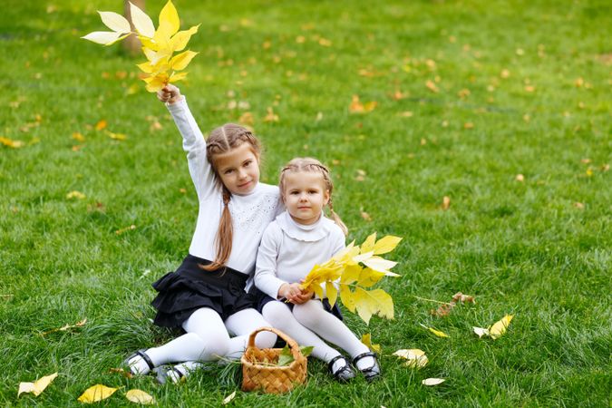 Two young blonde girls sitting in the park collecting leaves with basket