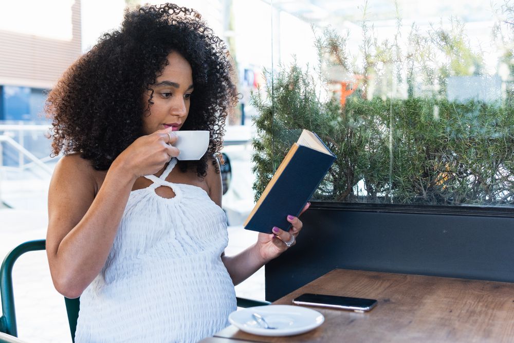 Pregnant Black woman reading book and drinking coffee