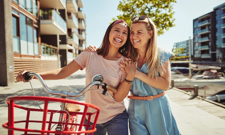 Two happy female friends laughing with arms on shoulders while walking with bike next to the river