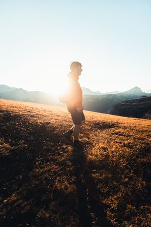 Side view of man in red jacket with backpack walking on shrubland at sunset