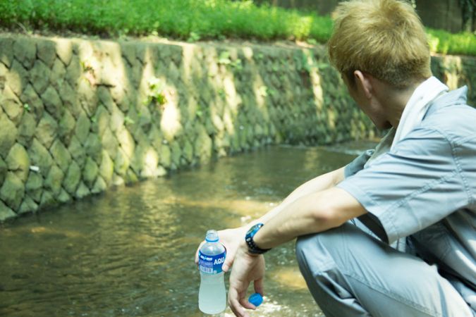 Man in work suit holding a cold bottle of water crouching beside stream river