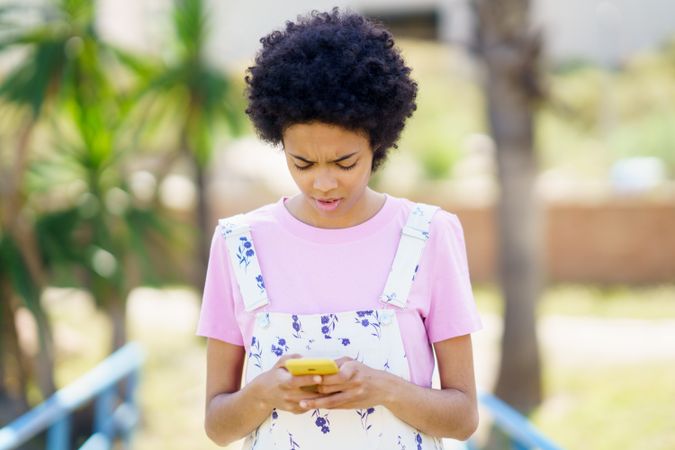 Confused female in floral overalls texting on phone outside