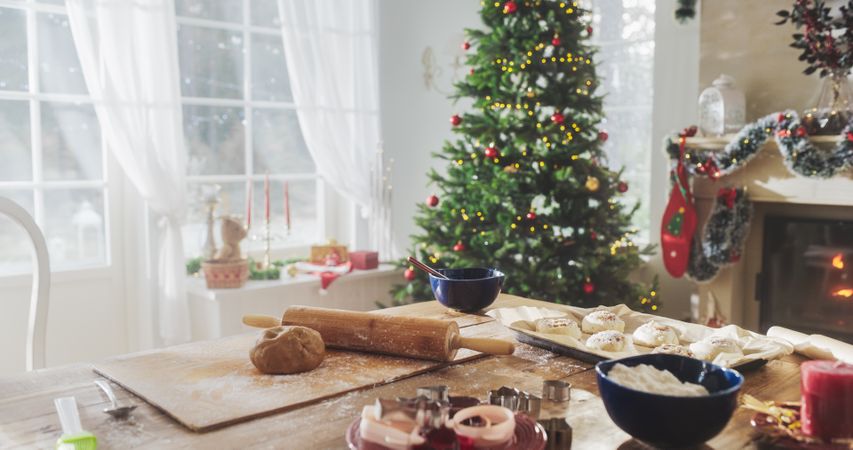 Peaceful Snowy Christmas Morning: Shot of a Table with Baking Ingredients and Utensils in a Decorated Corner of a Modern House with a Christmas Tree, Fireplace and Gifts.