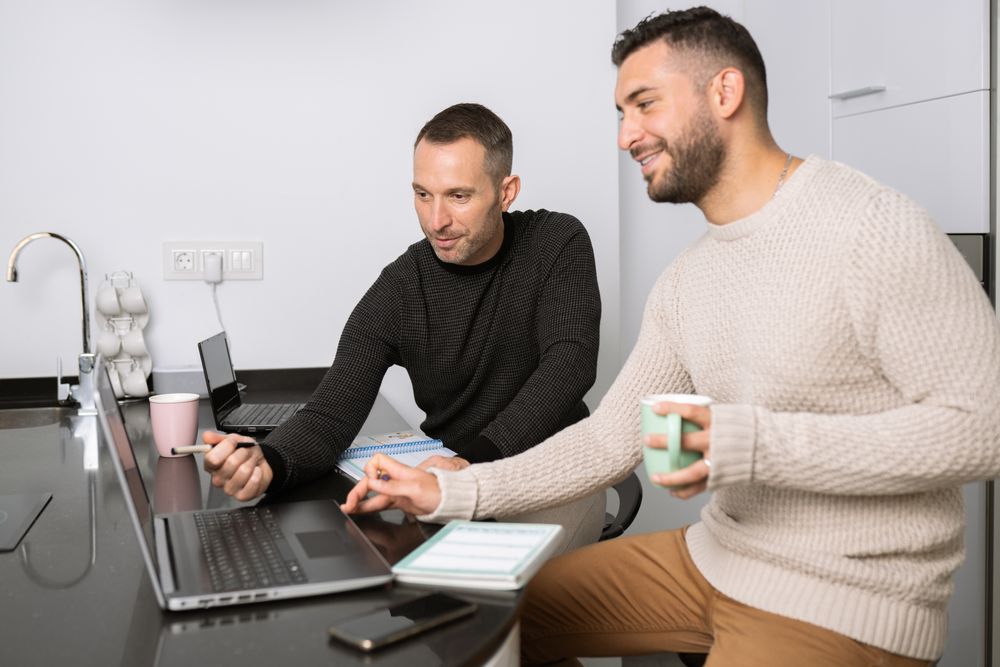 Two men sitting at kitchen counter and smiling at laptop