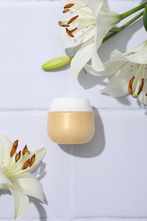 Jar with cream and lilies on light background, top view