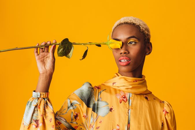 Black woman with short blonde hair holding yellow rose over eye