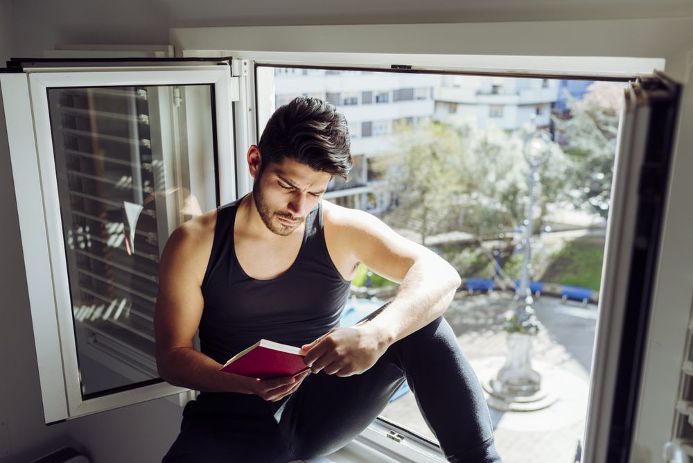 Young male sitting on window and reading book