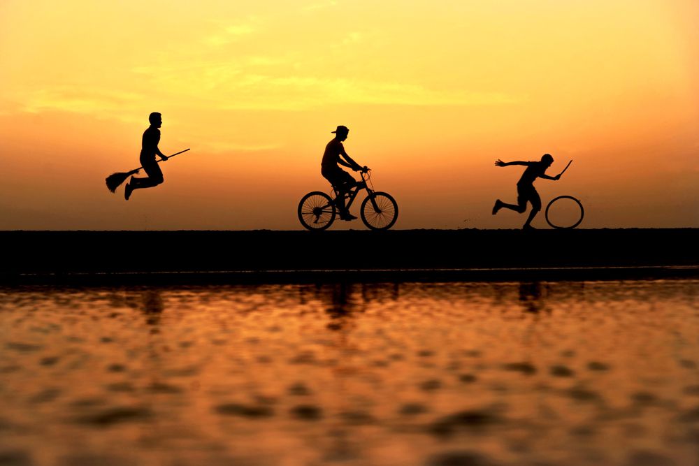 Silhouette of person riding witches broom and person riding a bicycle and another person moving a wheel near body of water during sunset
