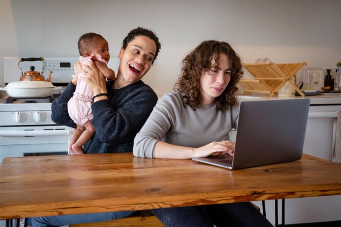 Woman on laptop in kitchen at home with partner and baby