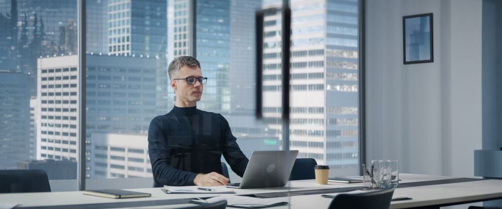 Businessman Sitting Down At His Desk And Starts Typing On Laptop Computer In Corner Office With Megapolis Cityview.