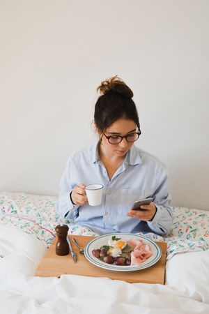 Young woman with breakfast on bed using smart phone