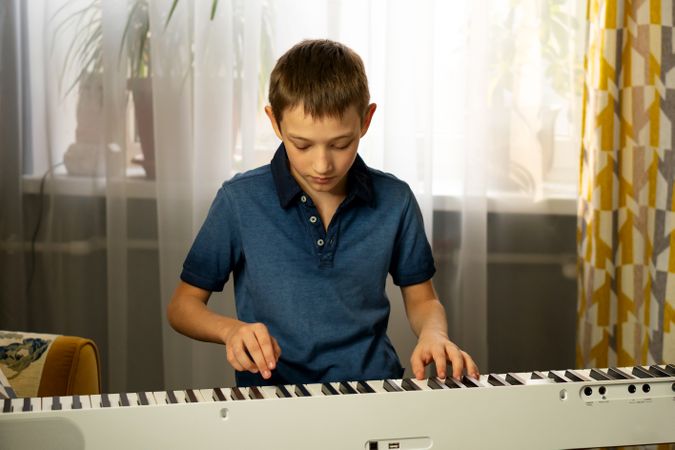 Boy in blue shirt playing piano near window with curtains.