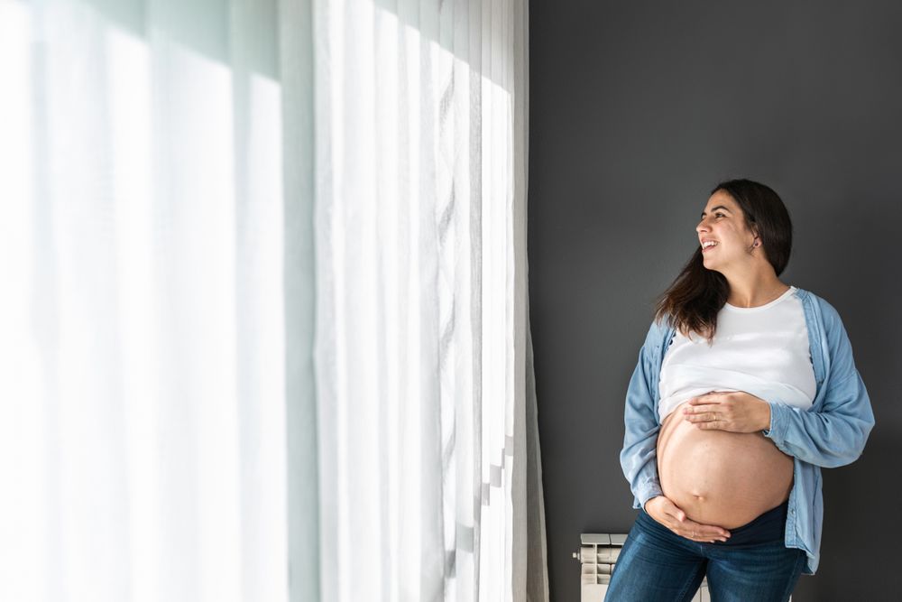Cheerful pregnant woman standing near window at home