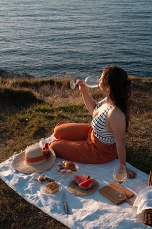 Woman with glass of drink on picnic mat looking at sea and mountains