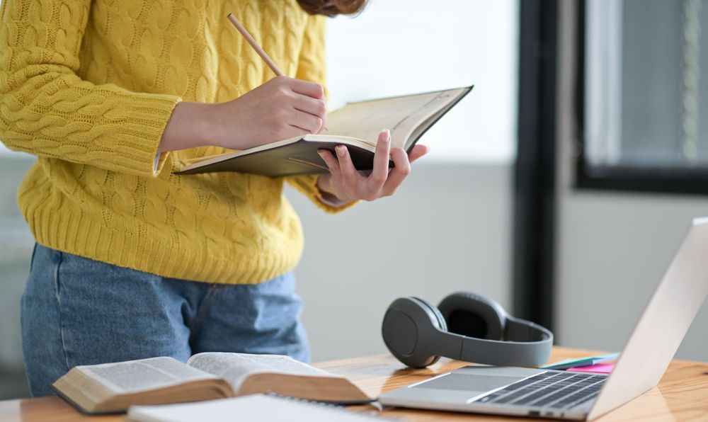 A female student takes notes in a notebook during an online class on a laptop from home.