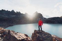 Back view of man in red jacket standing on rock near lake
