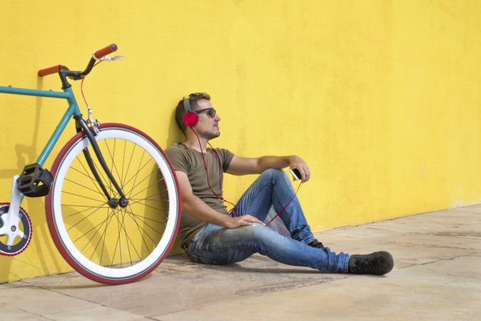 Male looking in distance in front of yellow wall next to bike and listening to music