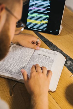 Back view of man reading a book beside a tablet computer