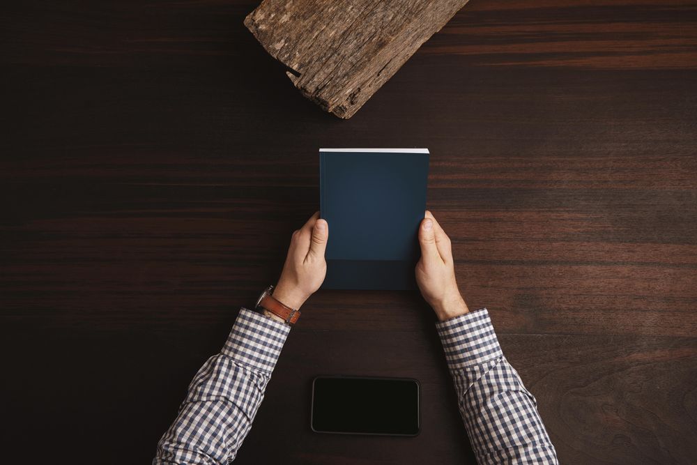 Arms in plaid shirt holding closed book with cell phone on table