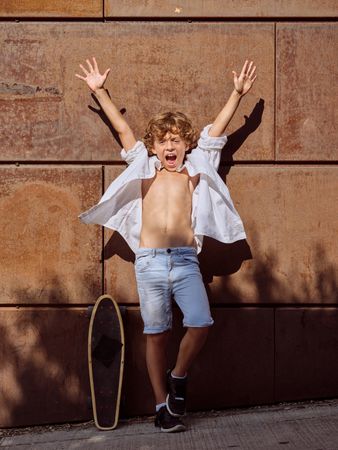 Excited boy with raised arms on street near skateboard