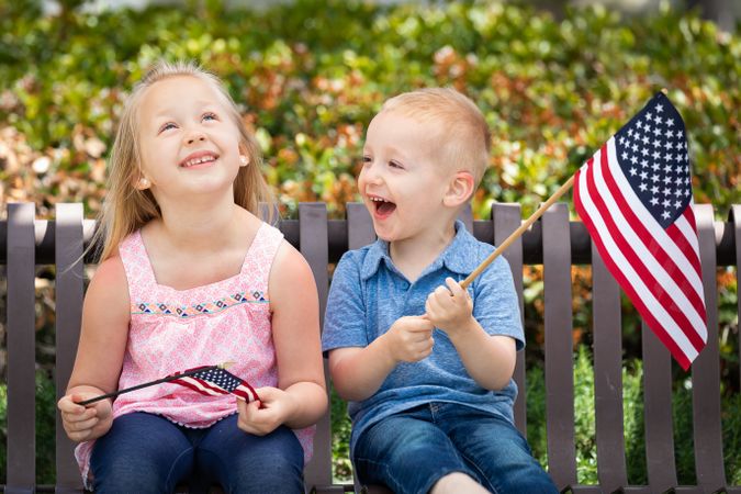 Young Sister and Brother Comparing Each Others American Flag Size On The Bench At The Park