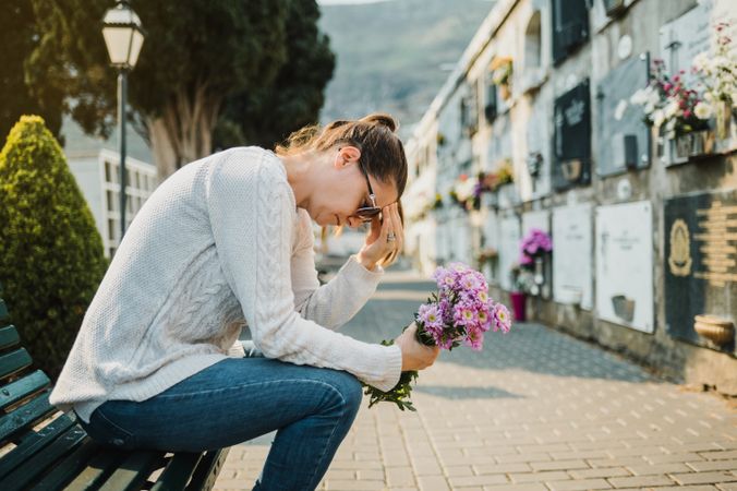 Sad woman crying sitting on bench with flowers in cemetery