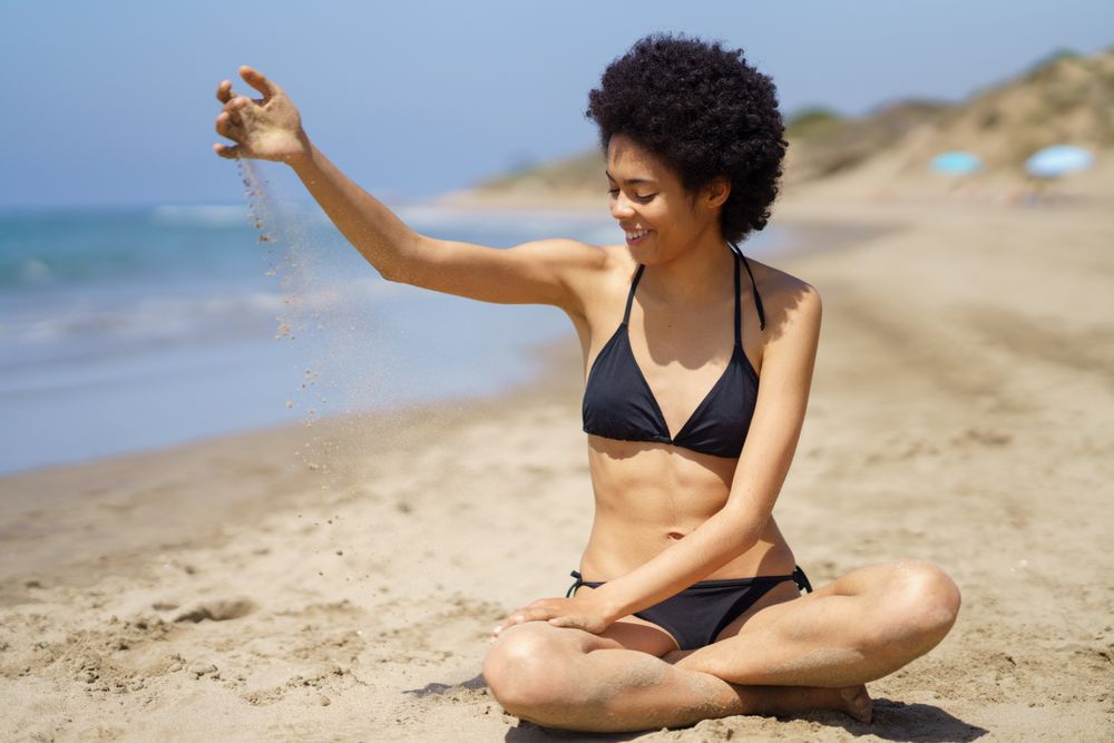 Female in bikini sitting crossed legged at the beach running sand through her hand