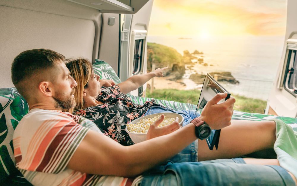 Male and female reclining on bed in motorhome enjoying the view with popcorn and digital tablet