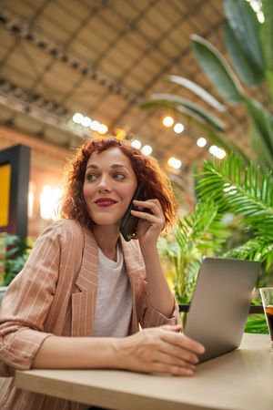 Cheerful young woman working on laptop in cafe and talking phone
