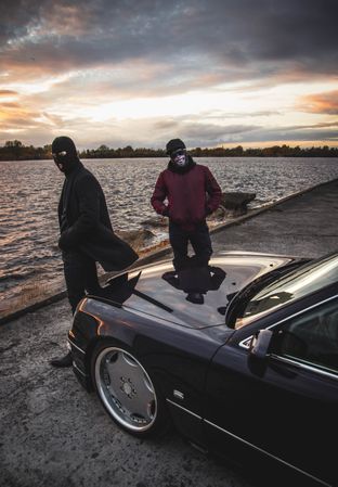 Two men with masks standing beside dark car by shoreline at sunset