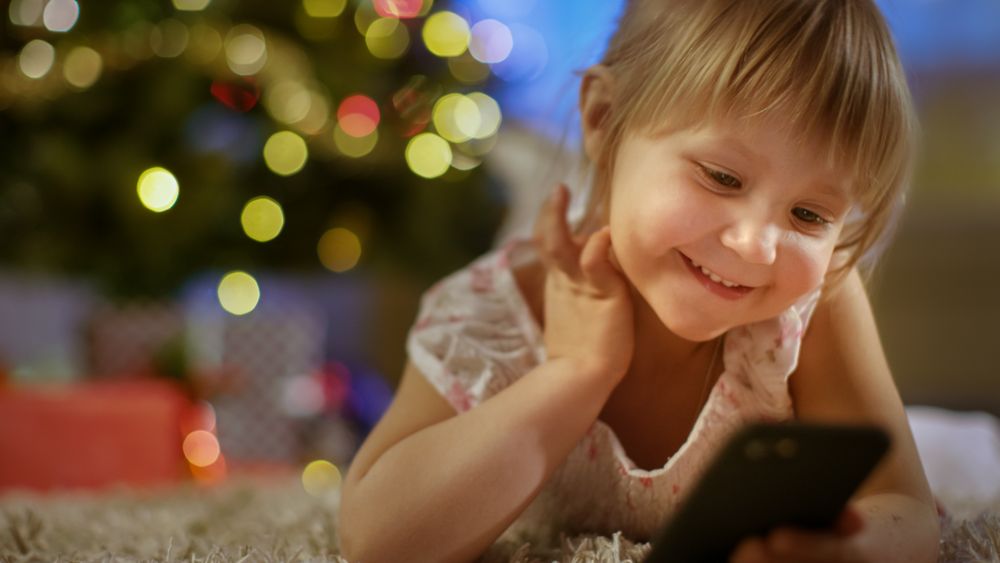 Little Girl With Smartphone in Her Hands Lies on the Carpet under the Christmas Tree.