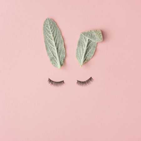 Bunny rabbit face made of natural green leaves with eyelashes on pastel pink background