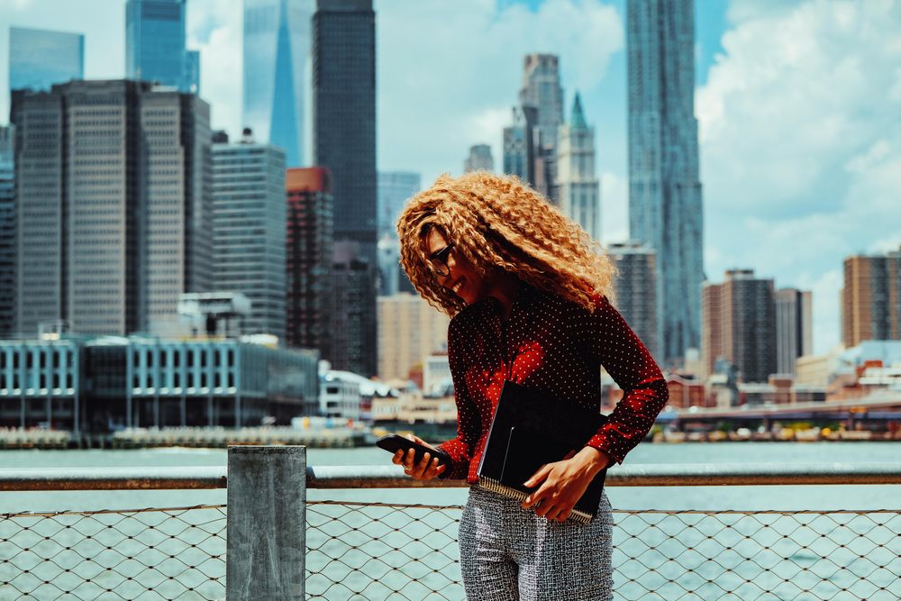 Happy professional Black female checking phone with Manhattan in the background