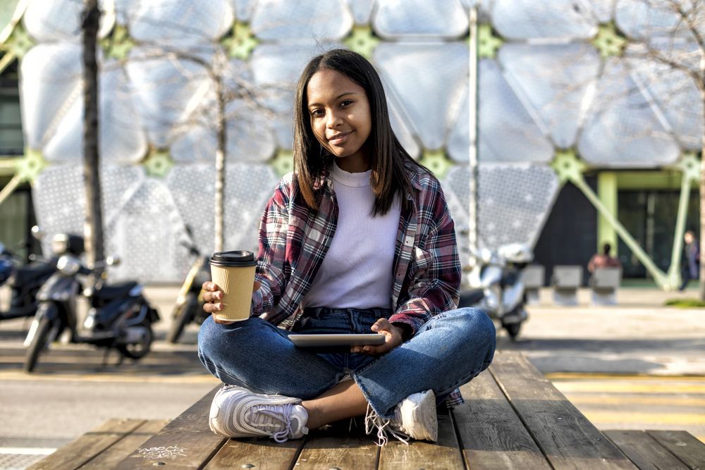 Female in plaid shirt sitting on table outside with tablet and to go drink