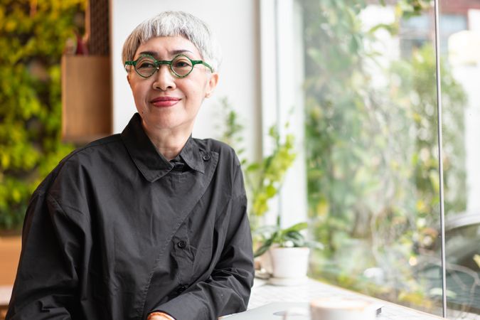 Grey haired Asian woman in trendy green glasses standing at window with coffee