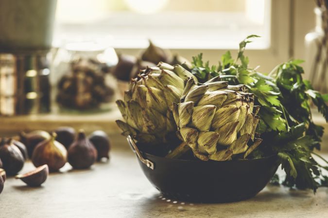 Whole artichokes in bowl with parsley on kitchen table with figs at window background with natural light.